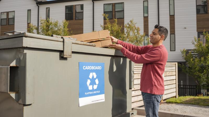 Man recycling flattened cardboard at apartment recycling dumpster