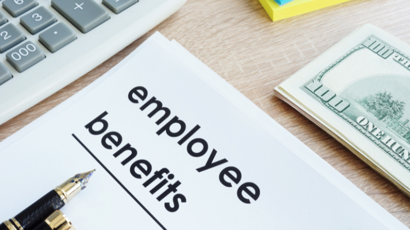 picture of a desk with a paper with the heading employee benefits, a computer, a pen and a hundred dollar bill