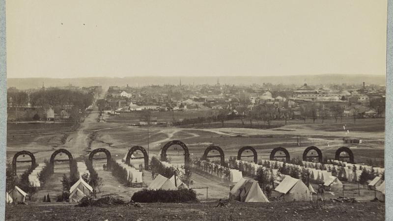 Photo taken from Shuter's Hill, the site of the George Washington Masonic National Memorial, looking east. Alexandria is visible in the background. The foreground shows lines of army tents with archways marking the roads between them. 