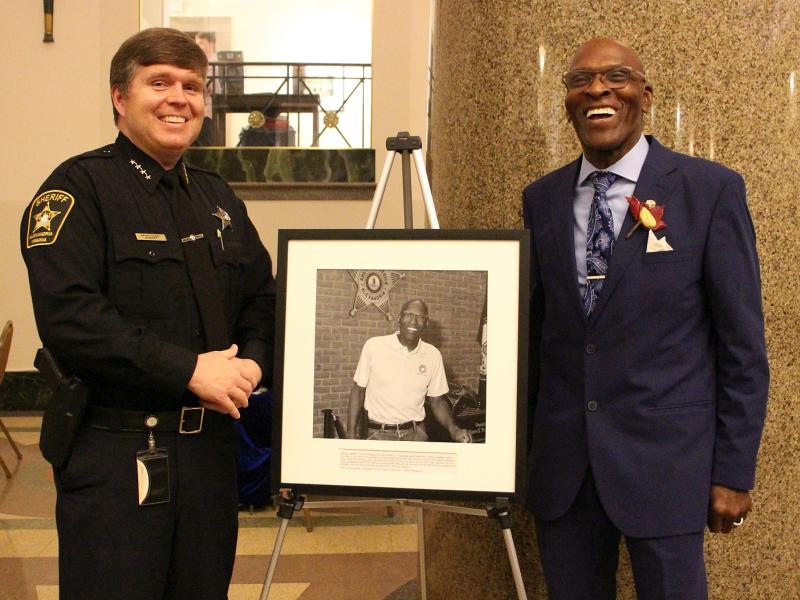 Sheriff in blue uniform and chaplain in blue suit standing and smiling with a photographic portrait of the chaplain on an easel between them