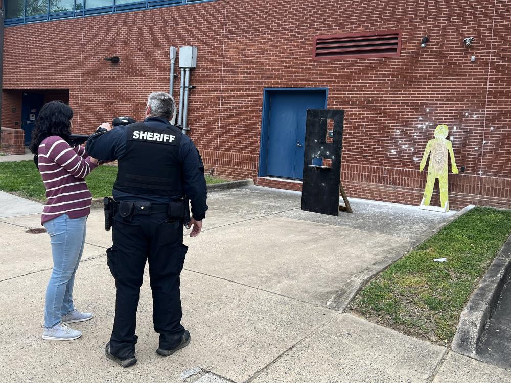 deputy showing civilian how to operate pepperball equipment with wooden silhouette in the background against brick wall