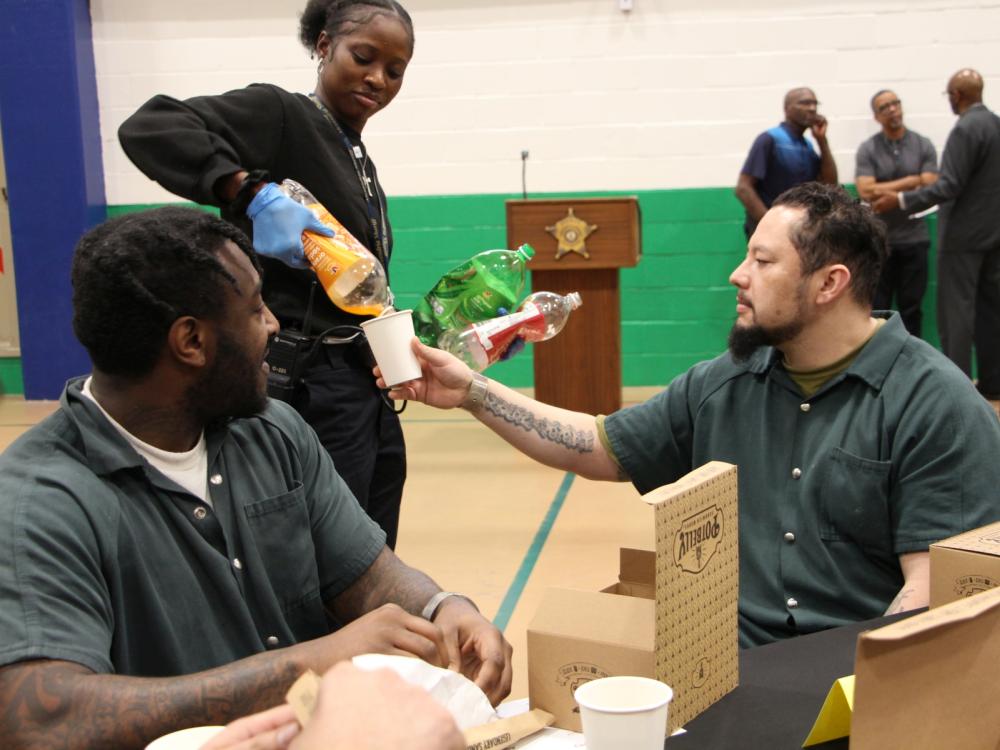 two inmates in green jumpsuits seated at a table as a recruit deputy pouring soft drink into one of their paper cups