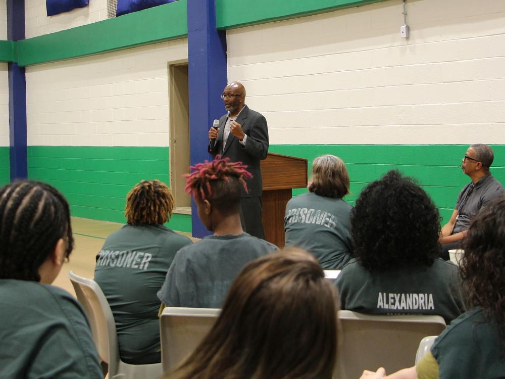 Man in a suit holding a microphone and speaking to several inmates who are seated at tables, wearing green jumpsuits and only visible from behind.