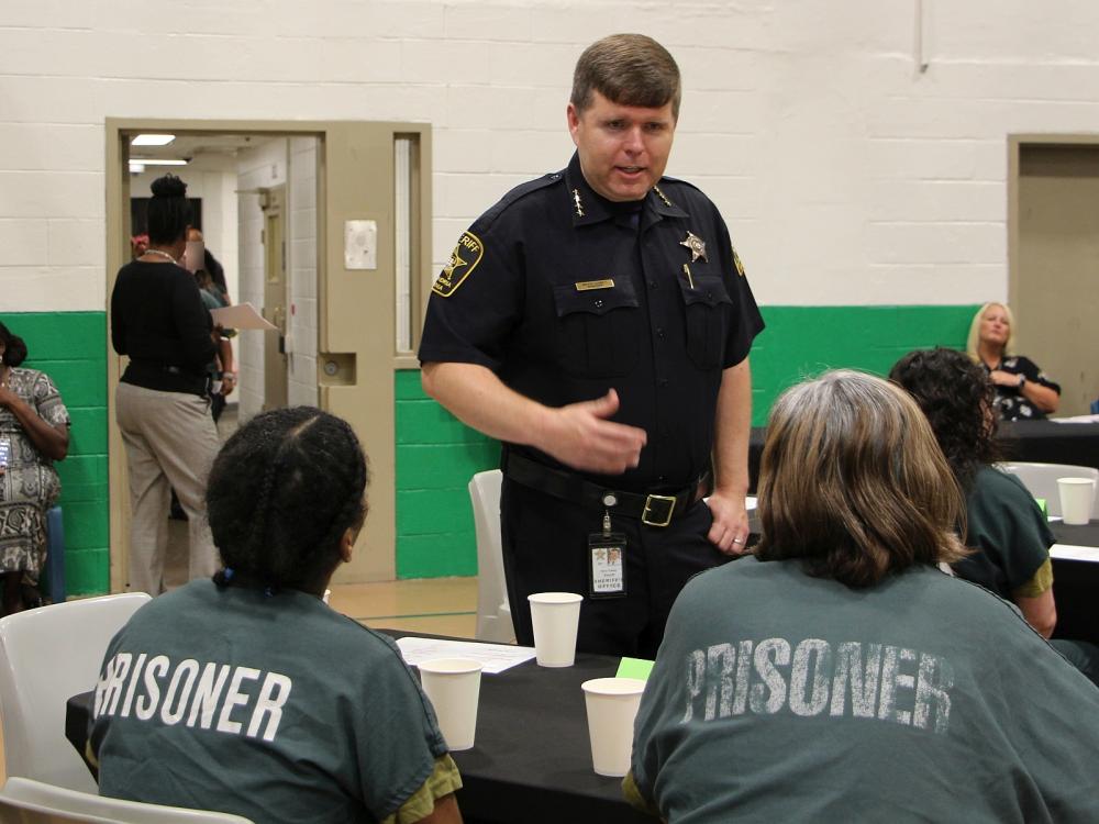 Sheriff talking with two inmates who are seated at a table and wearing green clothing that says "Prisoner" in white lettering on the back