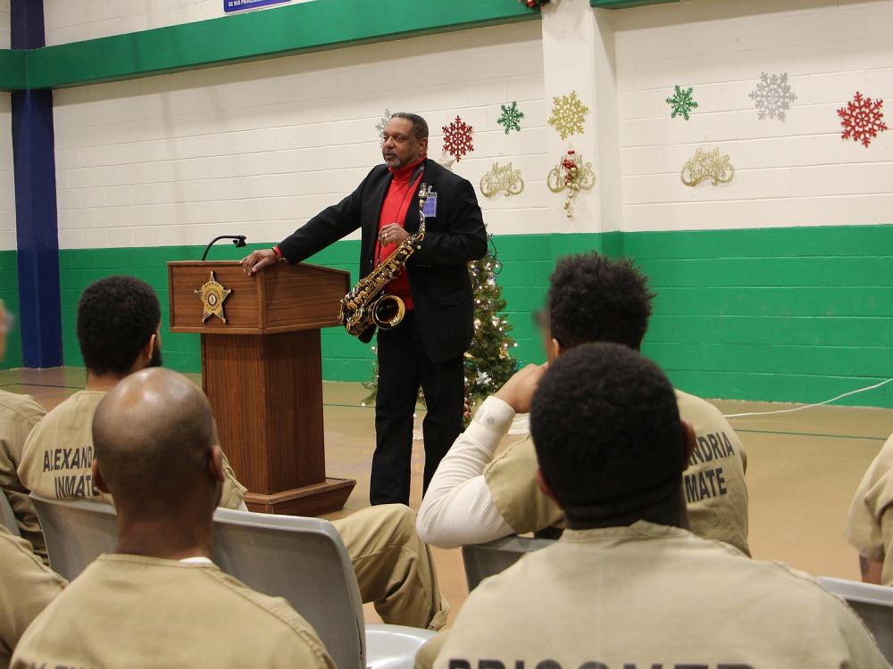 man wearing a black suit and red shirt and holding saxophone speaking to inmates wearing tan jumpsuits