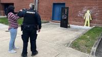 deputy showing civilian how to operate pepperball equipment with wooden silhouette in the background against brick wall