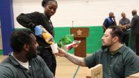 two inmates in green jumpsuits seated at a table as a recruit deputy pouring soft drink into one of their paper cups