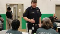 Sheriff talking with two inmates who are seated at a table and wearing green clothing that says "Prisoner" in white lettering on the back