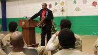 man wearing a black suit and red shirt and holding saxophone speaking to inmates wearing tan jumpsuits