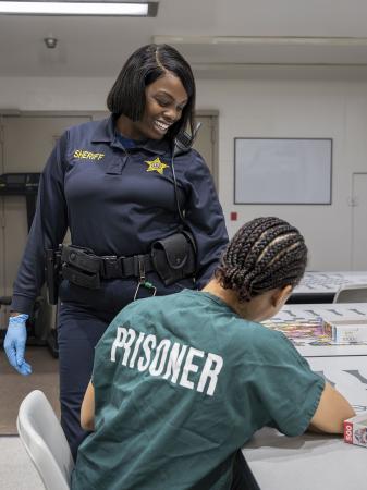 Deputy in blue uniform standing and smiling and engaging with an inmate seated and wearing a green jail uniform that says prisoner in white letters on the back