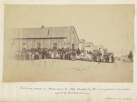 A photo showing approximately 100 children and a few adults standing in front of a long, low wooden building with windows, with frame buildings in the background