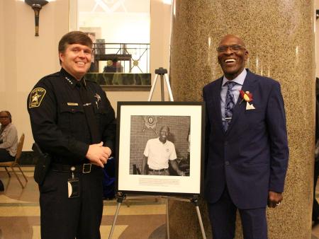 Sheriff in blue uniform and chaplain in blue suit standing and smiling with a photographic portrait of the chaplain on an easel between them